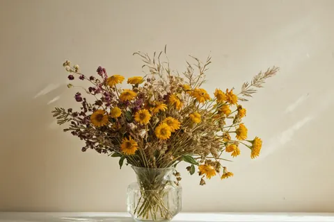 Dried yellow and purple flowers in a glass vase on a white surface with sunlight casting shadows on a beige wall.