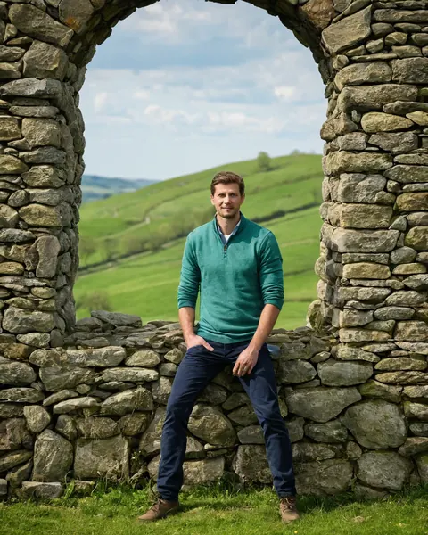 Man in teal sweater leaning on stone wall with green hills in background